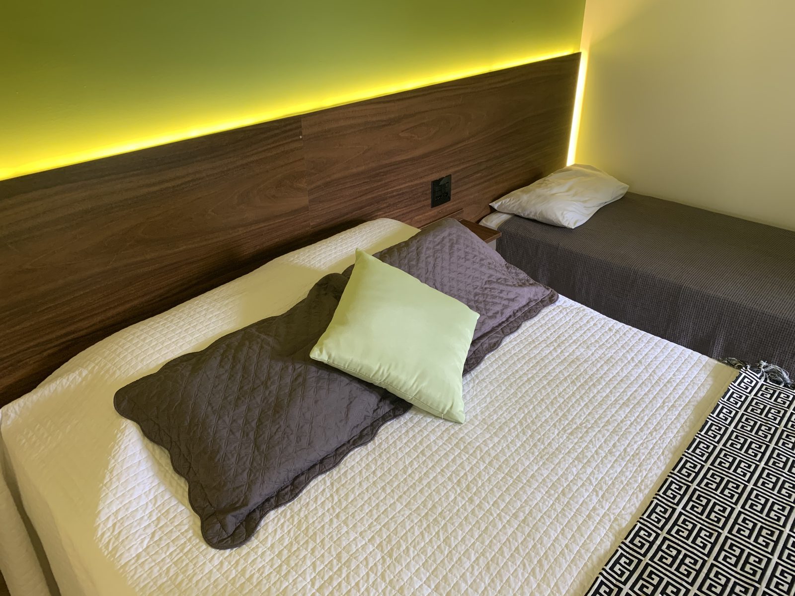 Detalhe da cama de casal com colcha branca e almofadas na Suíte Real, cabeceira de madeira com led