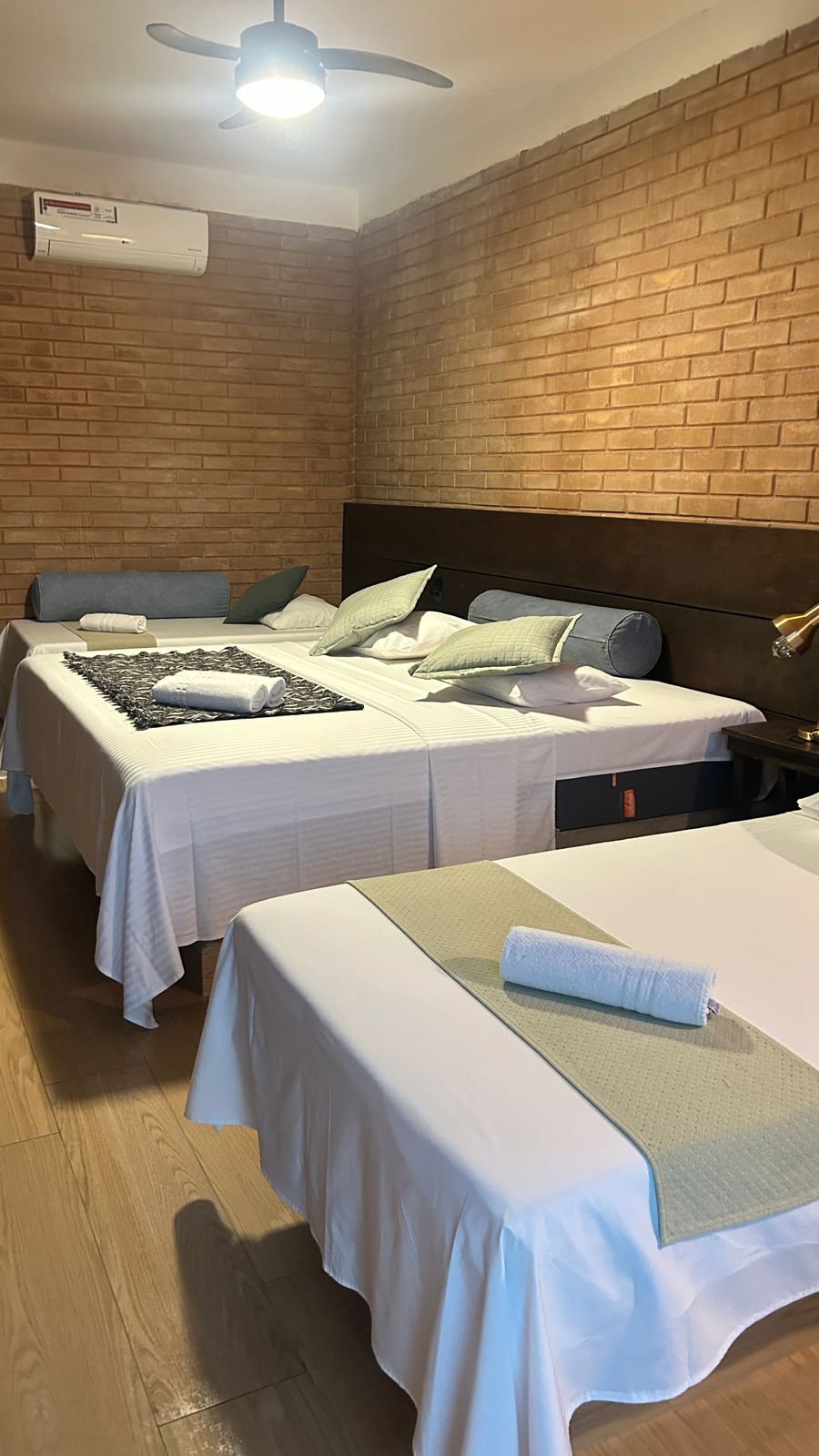 Suíte Família com três camas arrumadas, cabeceira de madeira, parede de tijolinhos e ventilador de teto no Hotel Holambra